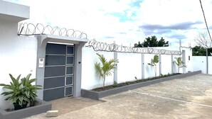 Un edificio con entrada protegida por una reja, camino de hormigón y pared blanca coronada con alambre de púas.