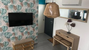 Un soggiorno moderno con TV a schermo piatto montata su una parete con carta da parati a motivi tropicali, una lampada pendente in fibre intrecciate, una panca in legno, un tavolino consolle in legno con lampade, e una porta sullo sfondo.