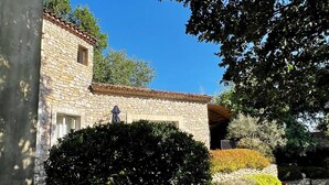 A stone building with a tiled roof, surrounded by well-maintained greenery and a clear blue sky.