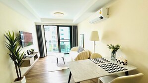 Een moderne hotelkamer met een houten vloer, een bed met een geruit kussen, een bureau met een schaakbord, een televisie, een plant en een balkon met uitzicht op gebouwen.