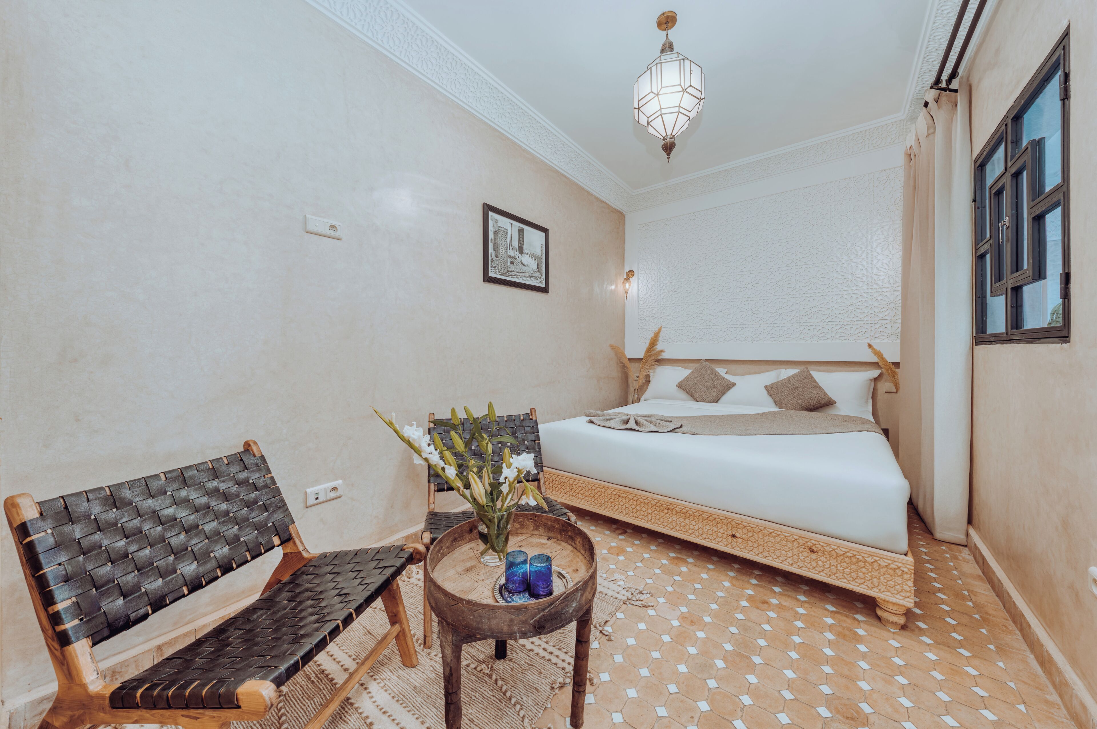 A room with a bed, a bench, a small table with a vase, and a hanging light fixture.