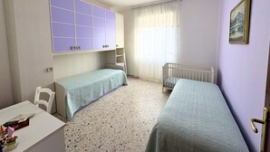 Una habitación pequeña y bien iluminada con dos camas individuales, un escritorio blanco y una ventana con cortinas translúcidas.