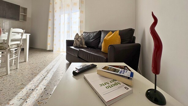 Una sala de estar con un sofá, una mesa de centro con un libro y un control remoto, un jarrón rojo, y una zona de cocina al fondo.