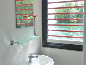 Un baño con un lavabo blanco, repisa de vidrio y ventana con persianas rojas.