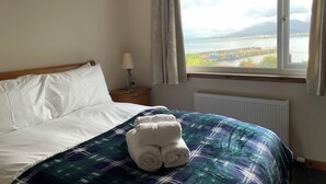 Una habitación de hotel con una cama, un ventanal con vistas al agua y montañas, una mesita de noche con lámpara, y una toalla enrollada sobre la cama.