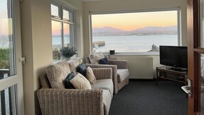 Una sala de estar acogedora con un conjunto de sofás de mimbre, televisor y vista al mar y las montañas al atardecer.