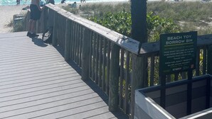 Ein Holzsteg führt zu einem Strand mit Palmen und einem Schild zum Ausleihen von Strandspielzeug.