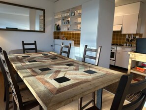A dining area with a long table, chairs, a kitchen with white cabinets, and a tiled backsplash.