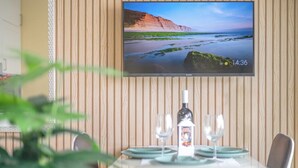 A dining area with a table set for two, a television displaying a scenic landscape, and a wooden wall in the background. - One bedroom and one living room apartments in Estoril Beach Villas (Estoril)