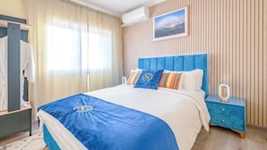 A hotel room with a large bed, a blue headboard, a nightstand with a lamp, and a window with curtains. - One bedroom and one living room apartments in Estoril Beach Villas (Estoril)