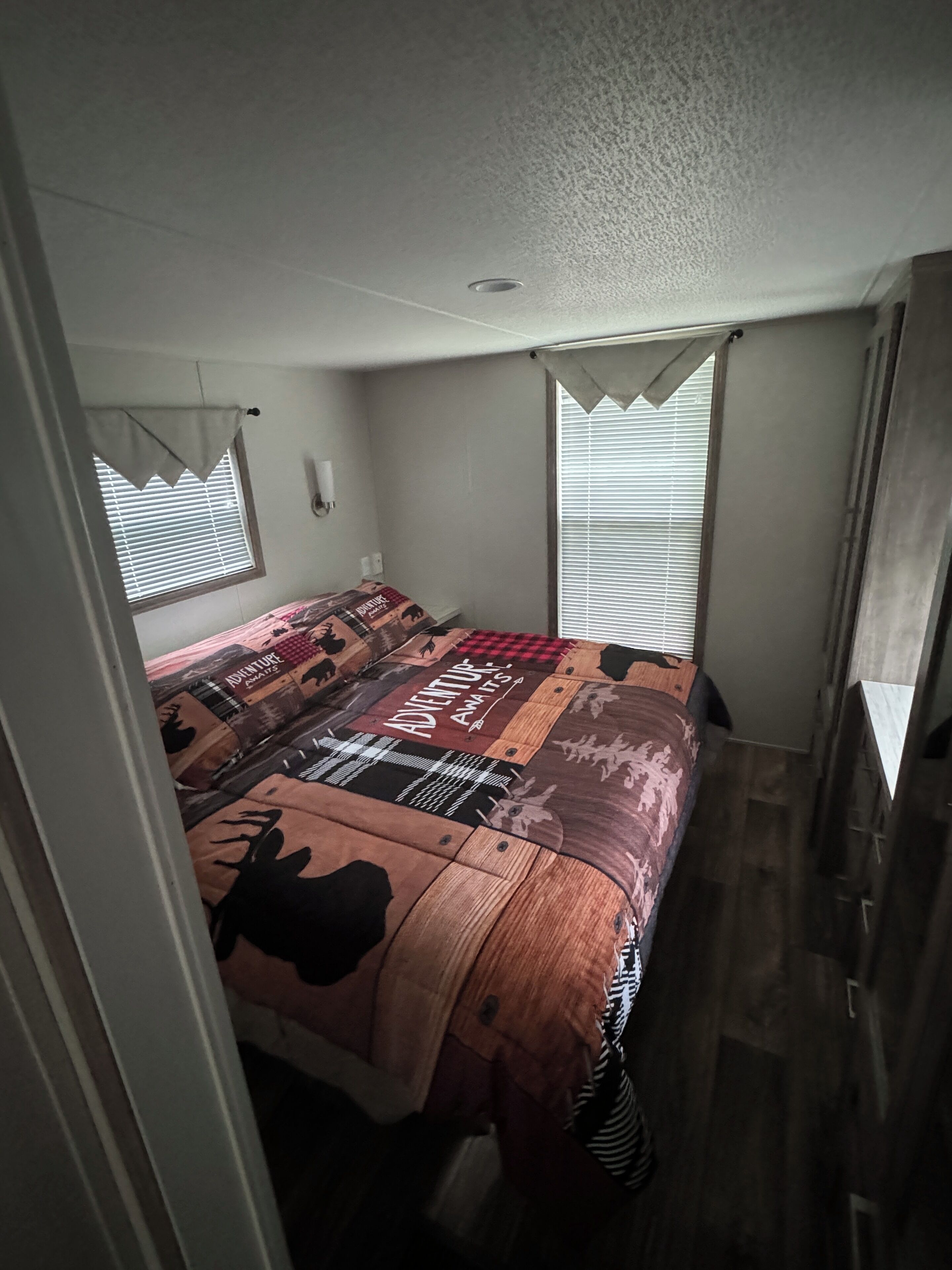 A bedroom with a bed covered in a patterned bedspread, a window with blinds, and a ceiling vent.