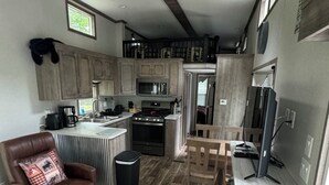 A modern kitchen with wooden cabinets, a built-in microwave, and a gas stove. There is a dining area with a wooden table and chairs, and a leather armchair. - Cozy 1 1/2-bedroom, 4 Bed Tiny House with Gas Heat/AC, WiFi ATV/Snowmobile (Park Falls)