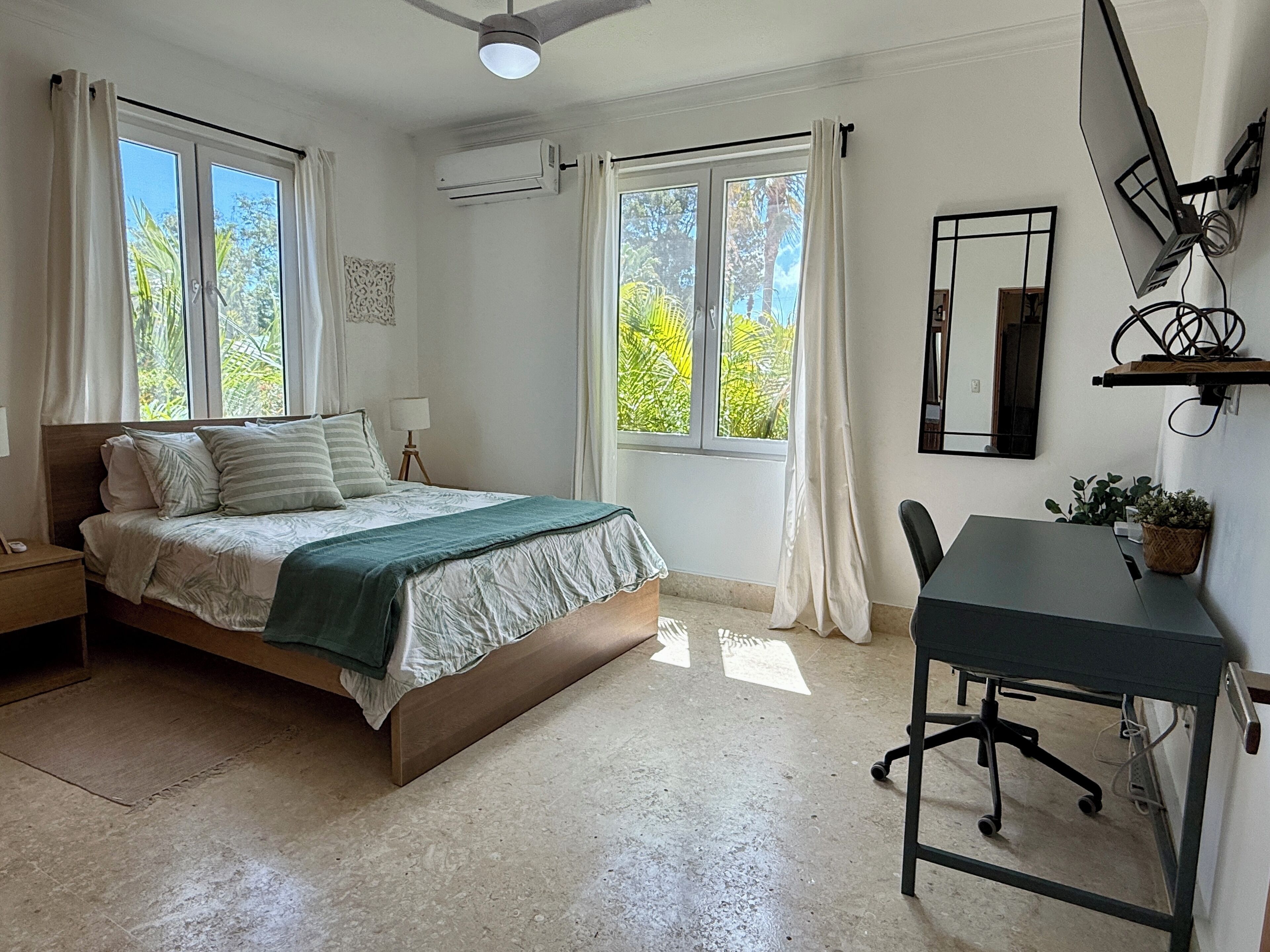 A bedroom with a bed, a desk, and a chair. There are windows with curtains, an air conditioning unit, and a ceiling fan.