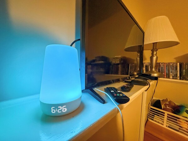 Une table de chevet avec une lampe de chevet blanche, une télécommande et un réveil numérique affichant 6 h 26.