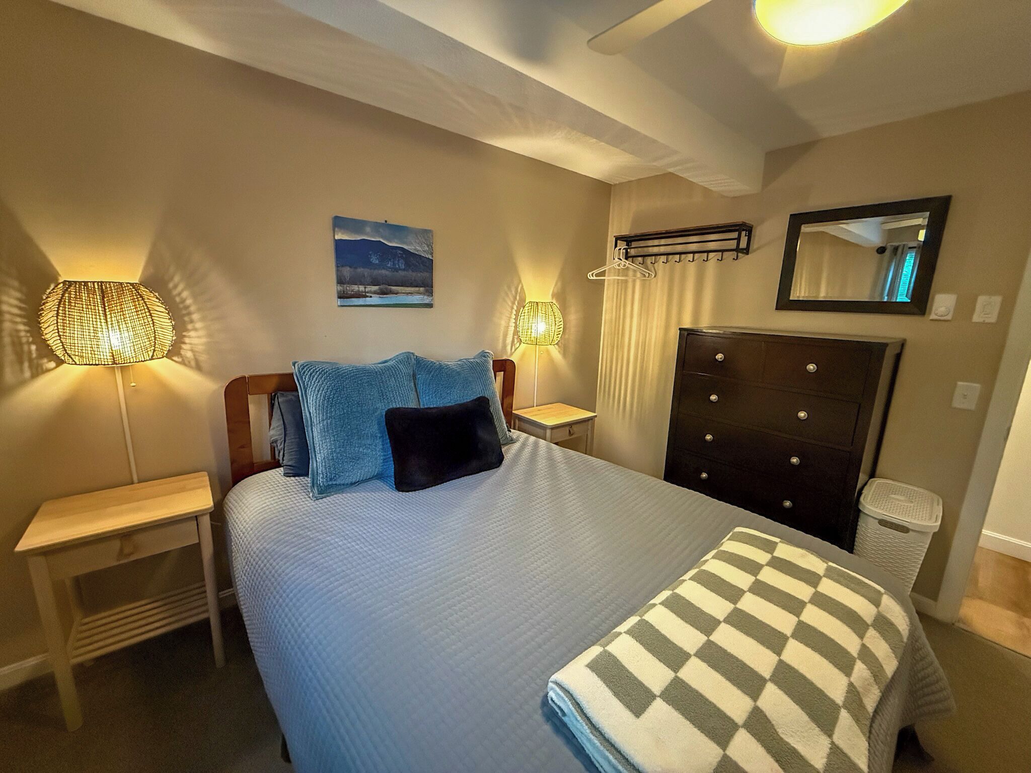 A neatly arranged hotel room with a bed, two bedside tables, a dresser, a hanging coat rack, a painting on the wall, and a mirror.
