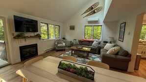 Een ruime woonkamer met een open haard, een eethoek met een plant in het midden, en een grote televisie aan de muur.
