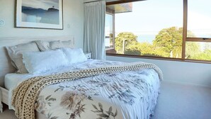 A bedroom with a large bed, a framed picture on the wall, and a view of trees and buildings through the window. - Views & Short Walk To Beach (Auckland)
