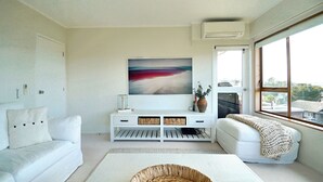 A modern living room with a white sofa, a white bench, a large painting, a white console table, and a large window with a view. - Views & Short Walk To Beach (Auckland)