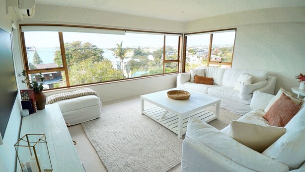 A modern living room with a white sofa, a coffee table, and a large window offering a view of a residential area. - Views & Short Walk To Beach (Auckland)
