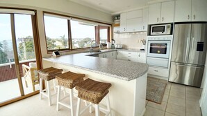 A modern kitchen with a large island, stainless steel appliances, and a view through the sliding glass doors. - Views & Short Walk To Beach (Auckland)