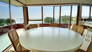 A round table with six wicker chairs in a room with large windows overlooking a body of water. - Views & Short Walk To Beach (Auckland)
