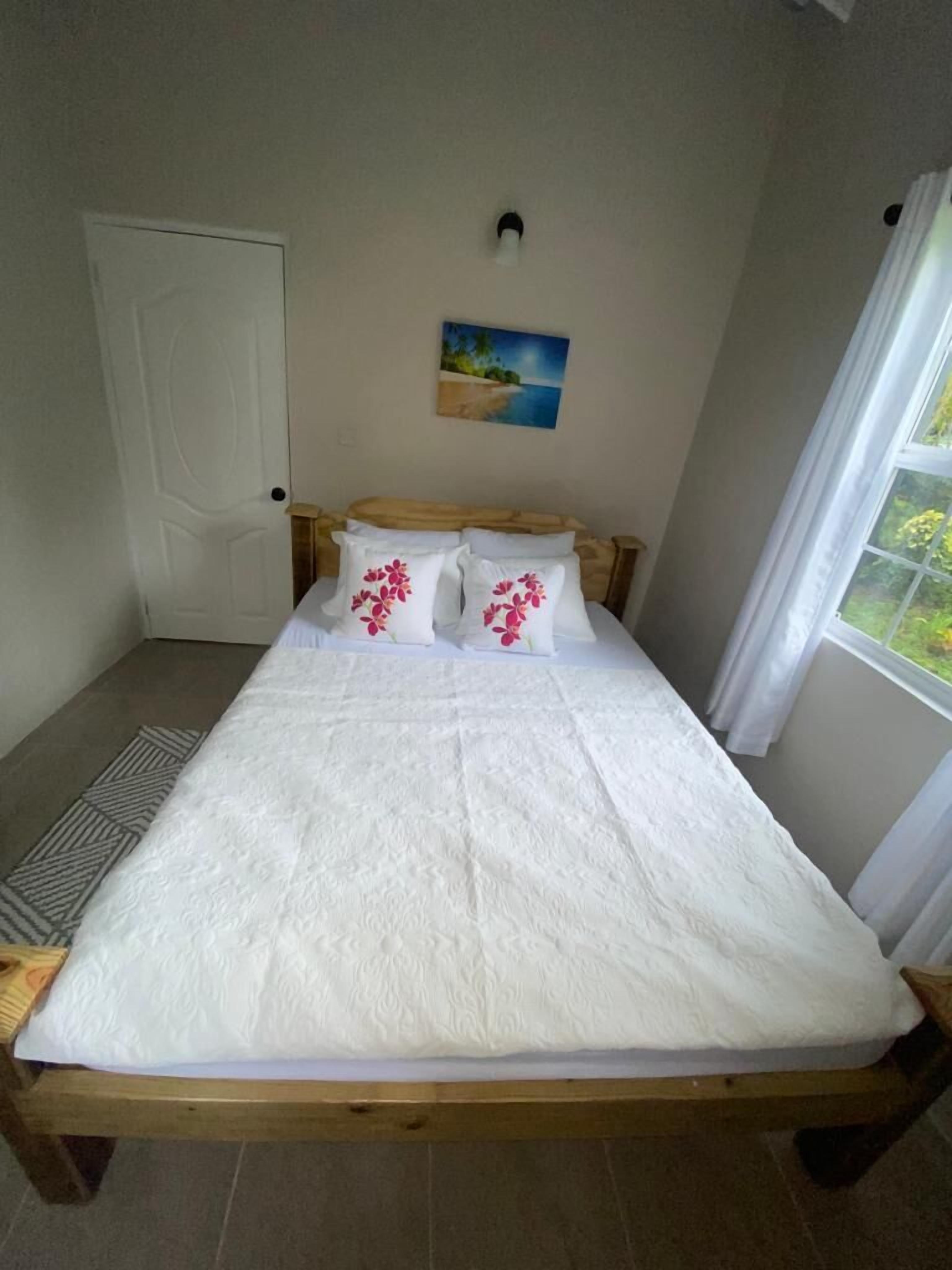 Una cama individual con estructura de madera, ropa de cama blanca y dos almohadas con diseños florales. Un cuadro de una escena de playa cuelga en la pared sobre la cama.