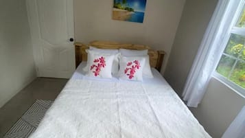 Una cama individual con estructura de madera, ropa de cama blanca y dos almohadas con diseños florales. Un cuadro de una escena de playa cuelga en la pared sobre la cama.