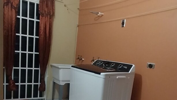 A room with a washing machine, a small table, and a window with curtains. - Charming 1-bedroom house with AC, WiFi in peaceful St. Helena Village Piarco (St helena village piarco)