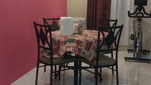 A dining area with a table set for four, surrounded by chairs. A white tablecloth covers the table, and a bottle of water is visible. The room has a red wall and a window with curtains. - Charming 1-bedroom house with AC, WiFi in peaceful St. Helena Village Piarco (St helena village piarco)