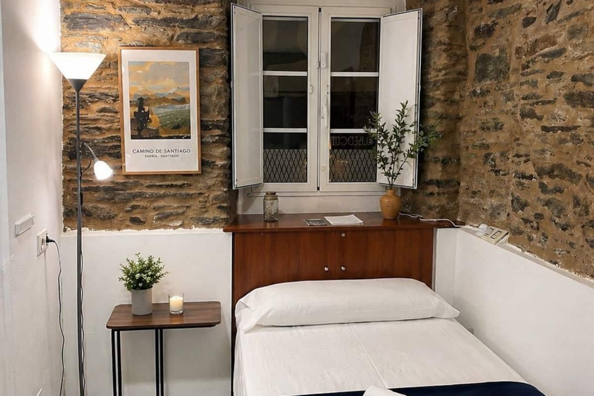 A small, cozy room with a stone wall, a bed with white bedding, a bedside table with a lamp, and a framed picture on the wall.