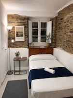 Una habitación pequeña y acogedora con pared de piedra, una cama con ropa blanca, una mesita de noche con lámpara y un cuadro en la pared.
