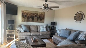 A living room with a brown leather sofa, a wooden coffee table, and patterned throw pillows. A ceiling fan is visible above.
