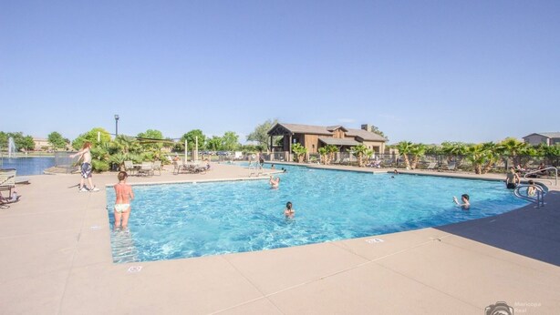 A swimming pool area with people swimming and relaxing. There are lounge chairs and umbrellas around the pool. - Lovely 3BR ranch home in Maricopa resort community w/ pools, courts, fitness ctr (Maricopa)