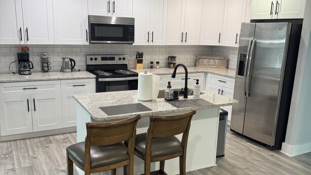 A modern kitchen with white cabinets, a stainless steel refrigerator, a microwave, and a stove. There is a dining area with two wooden chairs and a gray sofa. - Modern new townhouse near Stone Mountain. Sleeps 10. (Snellville)