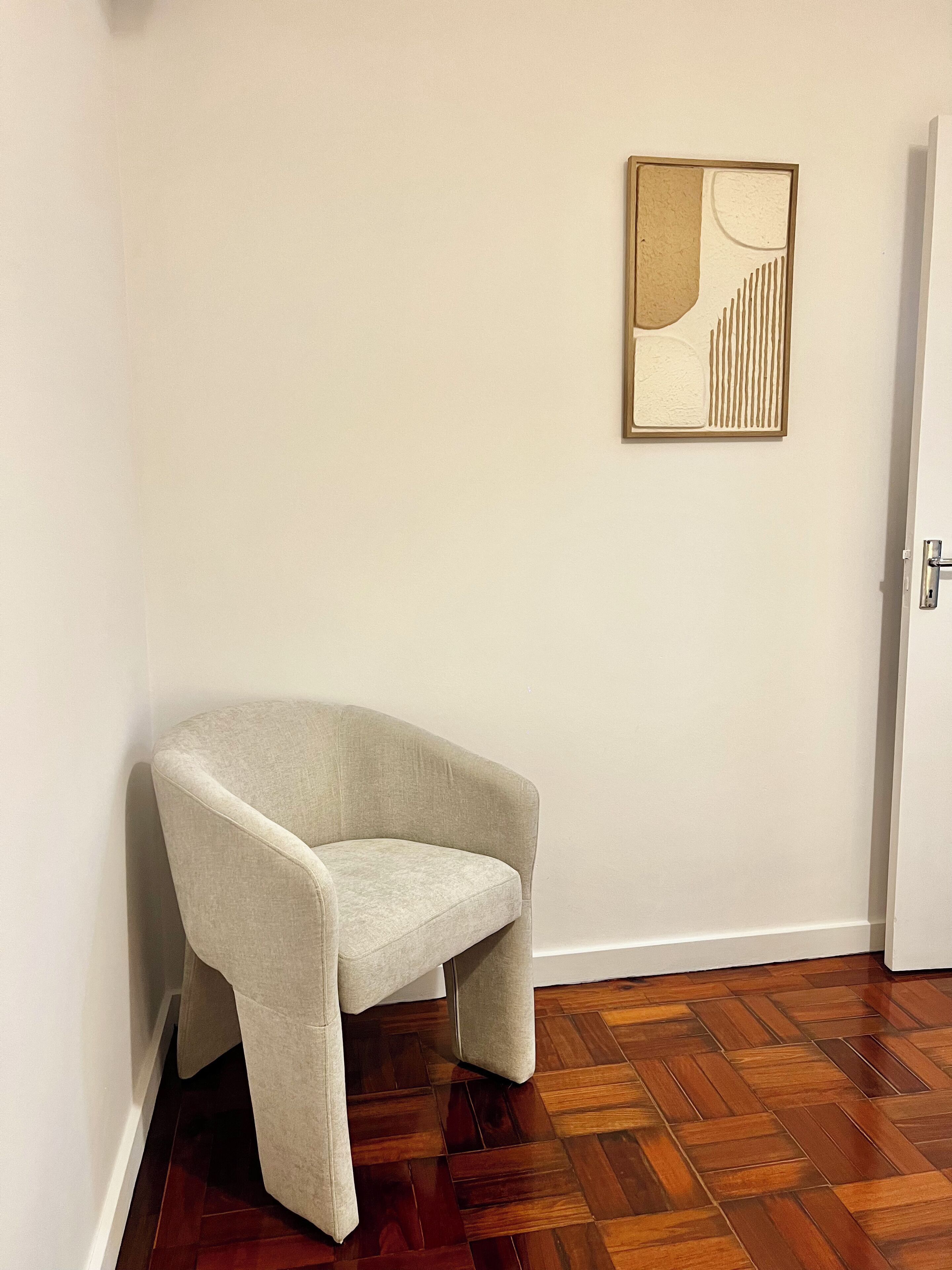 Ein Eckzimmer mit beigem Sessel, Holzdielen im Fischgrätmuster und einem gerahmten abstrakten Kunstwerk an der Wand.