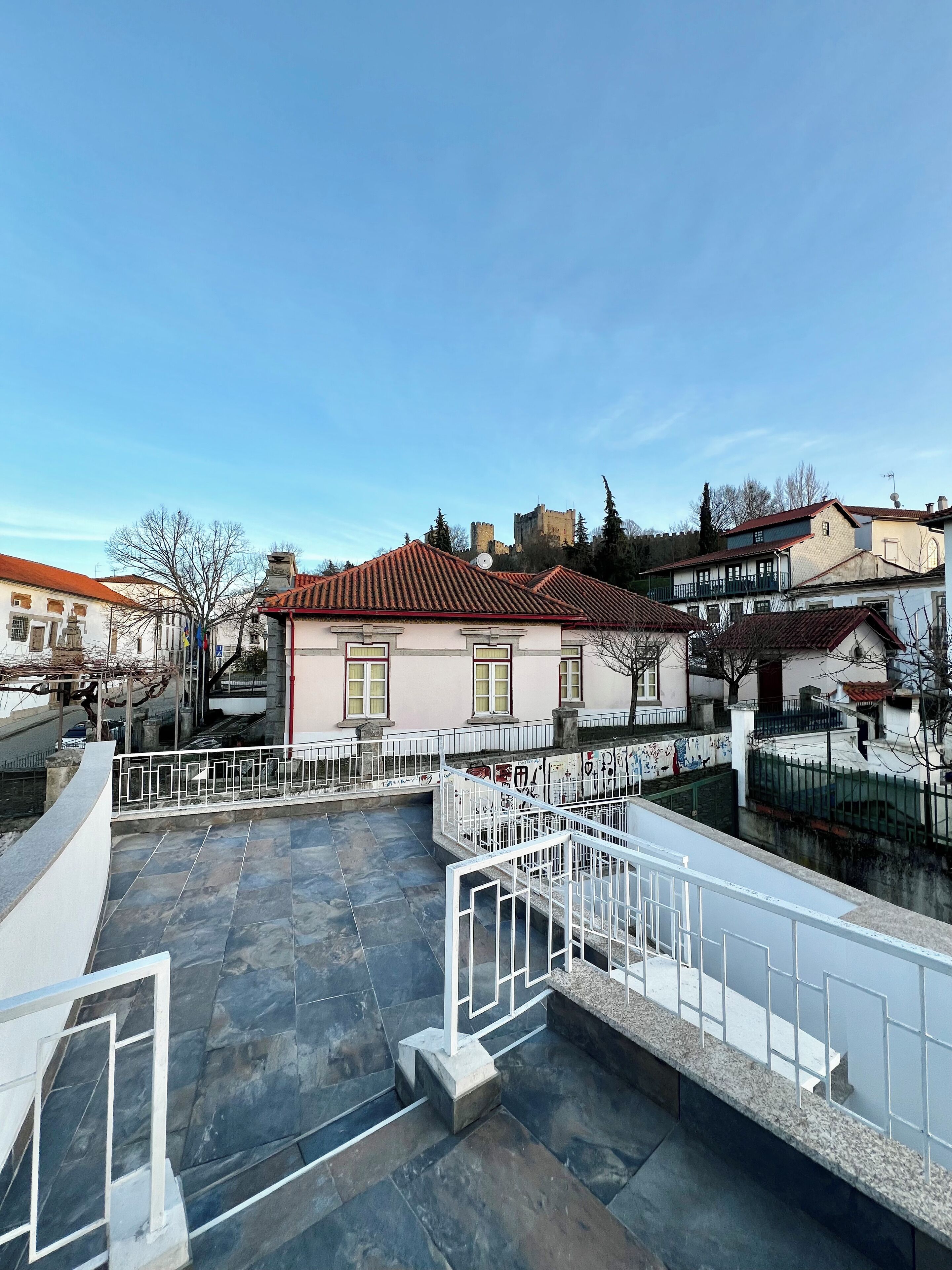 Una terraza en la azotea con suelo de piedra, barandillas blancas y vistas a edificios tradicionales y un castillo al fondo.