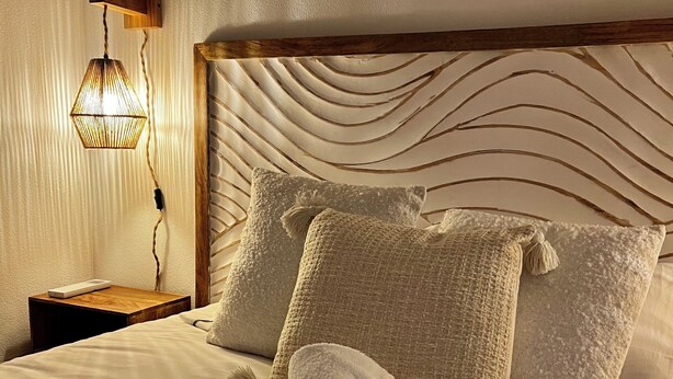 Una cama bien hecha con toallas enrolladas, un cabecero con textura y una lámpara de pared.