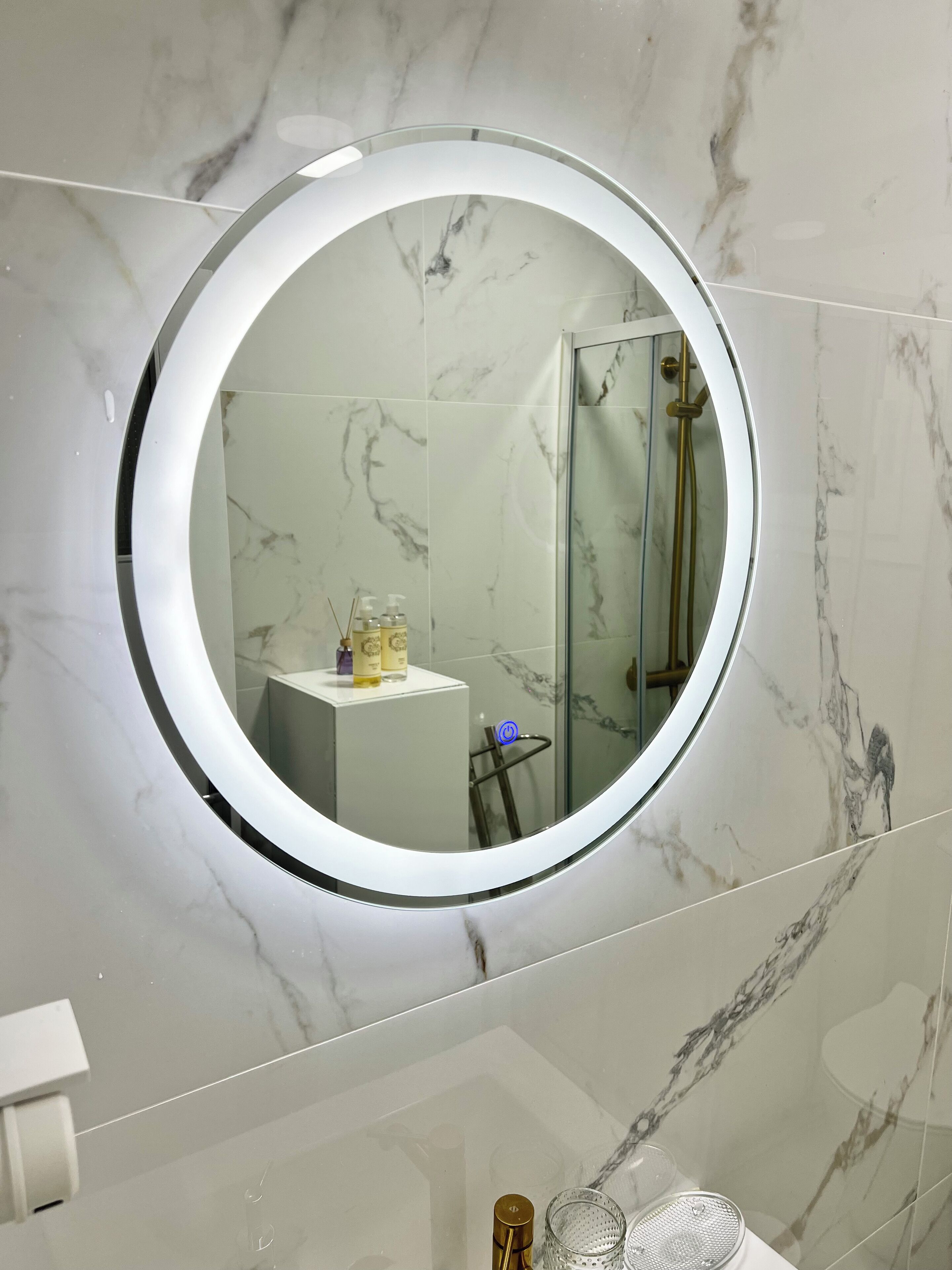 Un baño con un espejo redondo, baldosas de mármol, una cabina de ducha de cristal y una pequeña zona de lavabo con artículos de aseo.