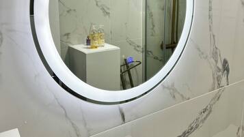 Un baño con un espejo redondo, baldosas de mármol, una cabina de ducha de cristal y una pequeña zona de lavabo con artículos de aseo.