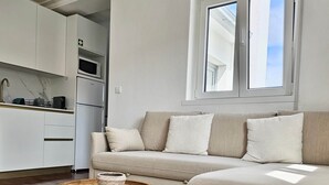 Una sala de estar moderna con un sofá color beige, una mesa de centro de madera y una cocina con armarios blancos y un microondas.