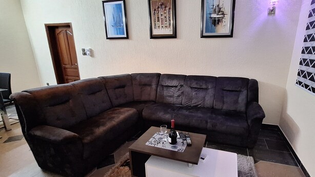 A living room with a dark brown sectional sofa, a small coffee table with a bottle and glass, and three framed artworks on the wall. - Room-deluxe 2 Bedrooms Apartment In Coto (Cotonou)