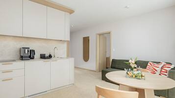 A modern kitchen with white cabinets, a coffee machine, and a sink. There is a dining area with a round table and chairs, and a green sofa with a floral arrangement.