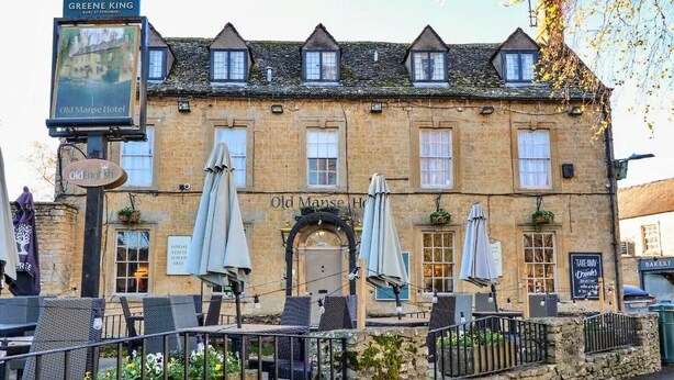 A traditional stone building with a sign reading "Old Mansse Hotel." There are outdoor tables with umbrellas and a stone wall in front.