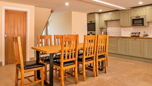 A dining area with a wooden table and chairs, a kitchen with light green cabinets and appliances, and a door in the background. - Little Rissington - Barn Six (Little Rissington)