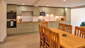 A modern kitchen with a dining area, featuring light green cabinets, a built-in oven, a microwave, and a wooden dining table with chairs. - Little Rissington - Barn Six (Little Rissington)