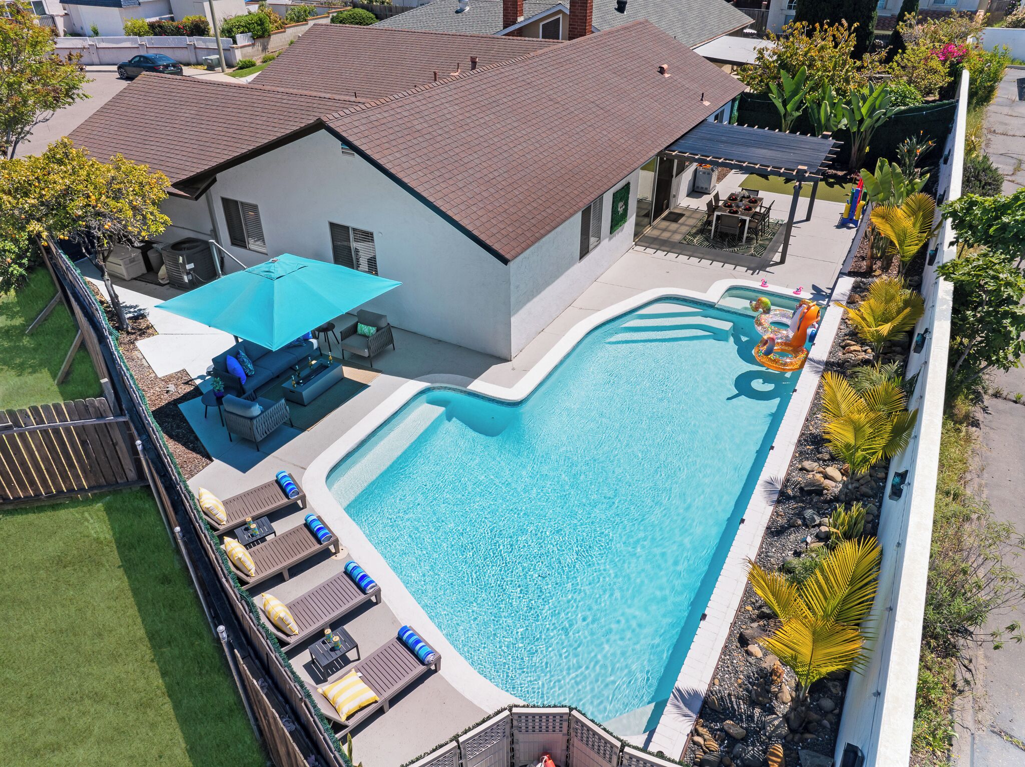 A residential swimming pool with a sun deck, lounge chairs, and a covered patio area.