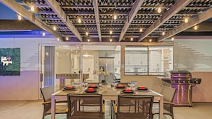 Un área de comedor exterior con una mesa preparada para cuatro personas, rodeada de sillas, una parrilla y vistas de la cocina.