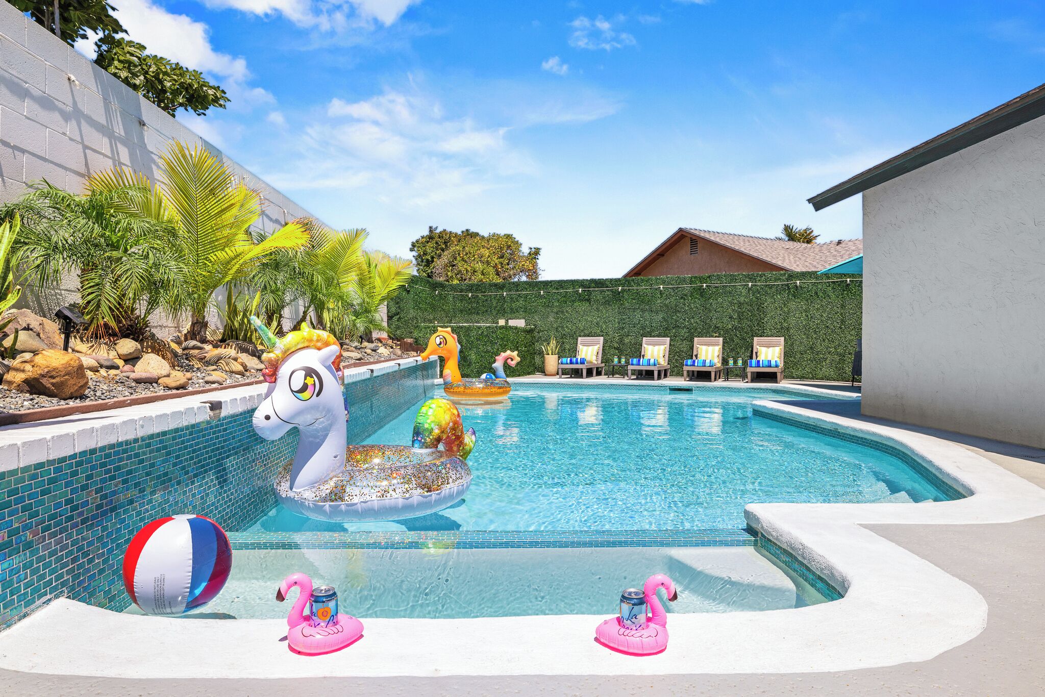 Une piscine agrémentée d’éléments décoratifs, comme un flotteur en forme de licorne et des flamants roses, entourée de chaises longues et d’un espace paysager avec des palmiers.