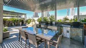An outdoor kitchen area with a glass-top table and wicker chairs, surrounded by stone walls and potted plants. - Luxury Apartment - Oracle Broadbeach  (Broadbeach)