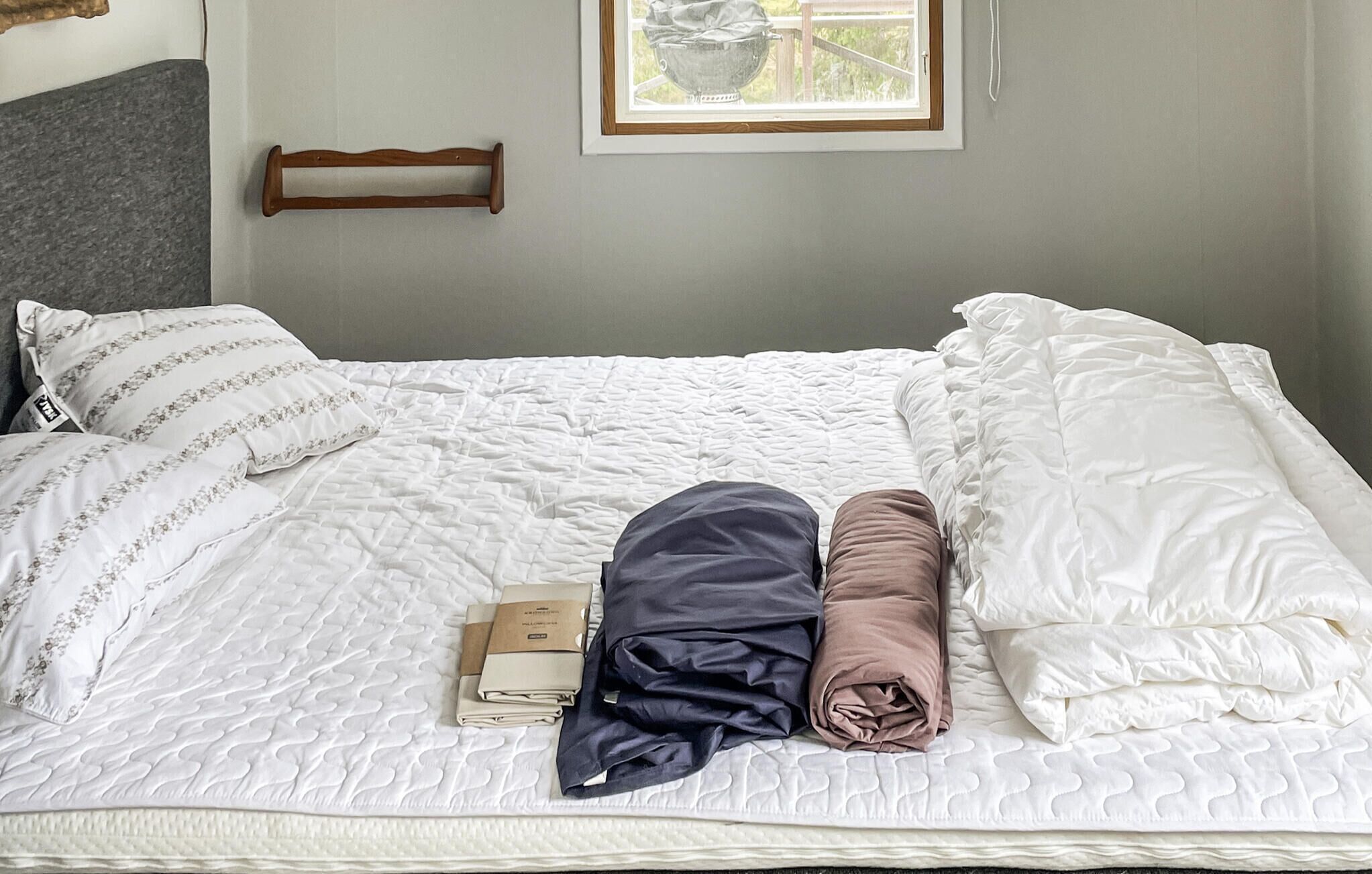 A neatly made bed with white bedding and pillows, a wooden headboard, and a window with a view of greenery.